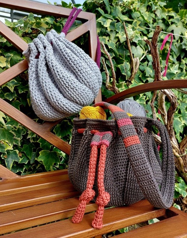 Gehäkelte Beuteltaschen in Hellgrau und Anthrazit auf Holzbank mit Garnknäueln.