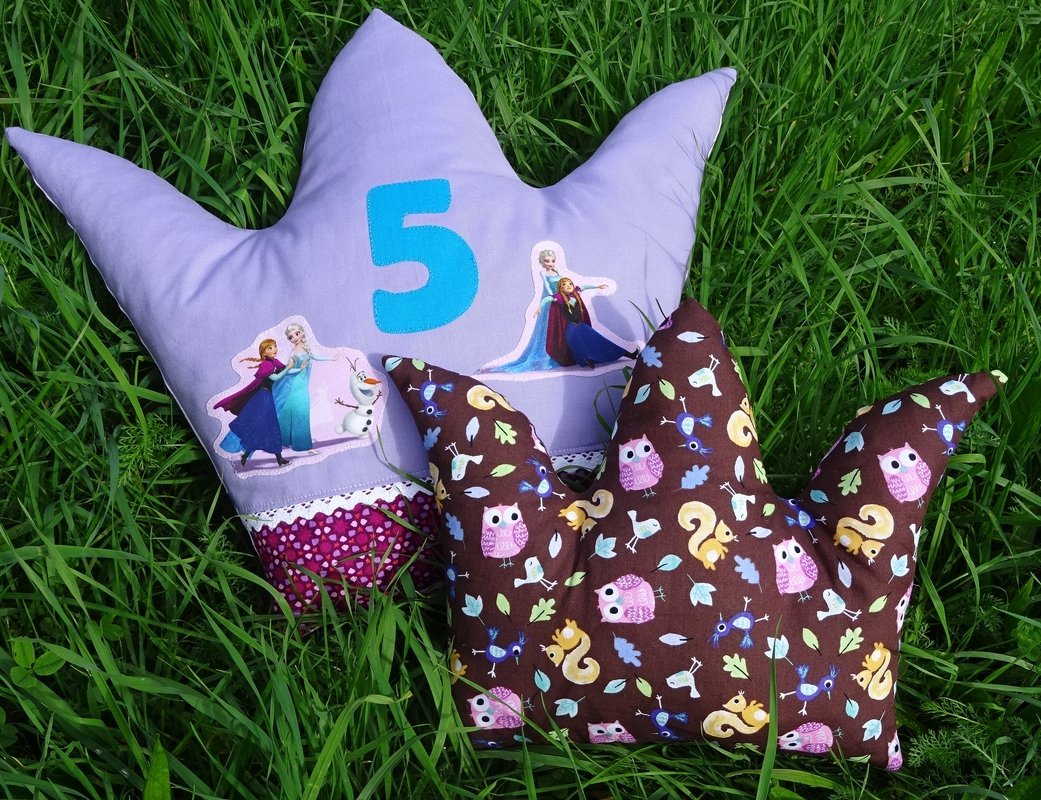 Zwei kronenförmige Kissen im Gras: lavendelfarbenes Kissen mit Zahl „5“ und Figurenaufnähern, braunes Kissen mit Eulen- und Eichhörnchenmuster
