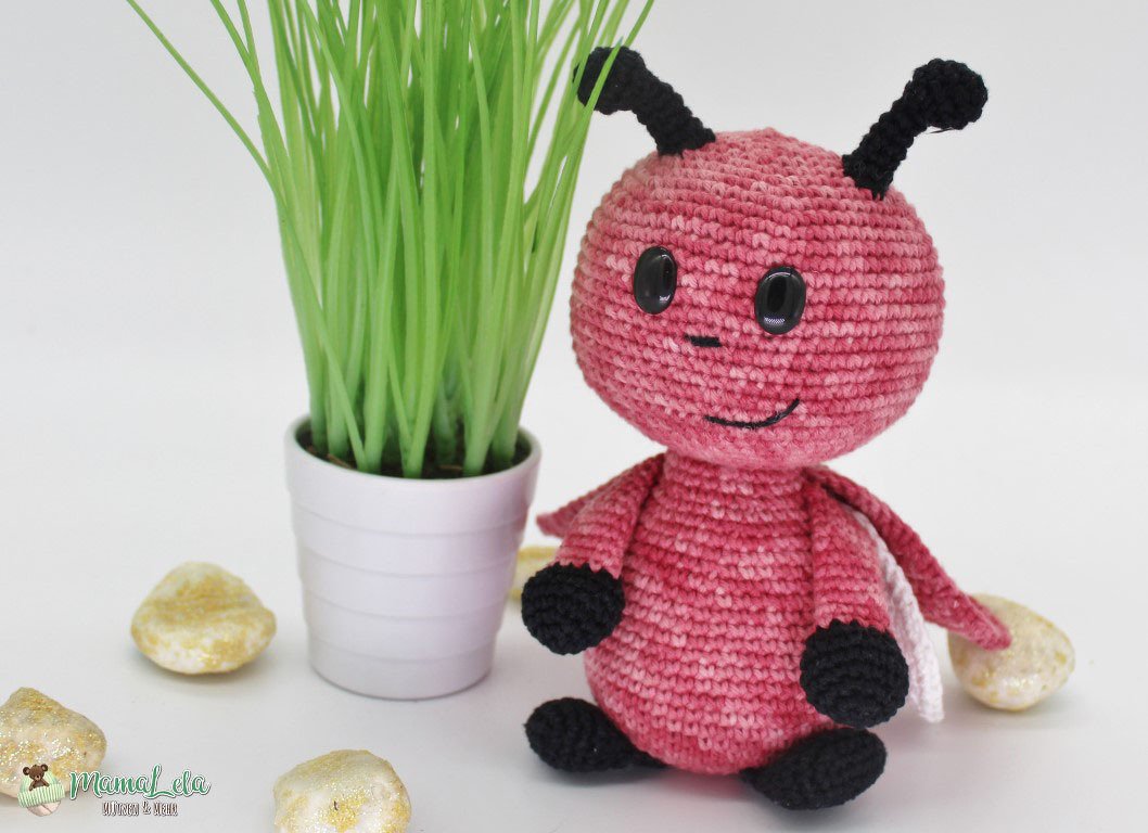 Rosa gehäkeltes Amigurumi-Käferchen mit schwarzen Antennen neben einem kleinen Topf mit Gras