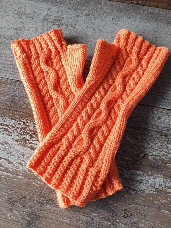 Nahaufnahme eines Paares orangefarbener, gestrickter Armstulpen mit Zopfmuster auf Holzuntergrund.
