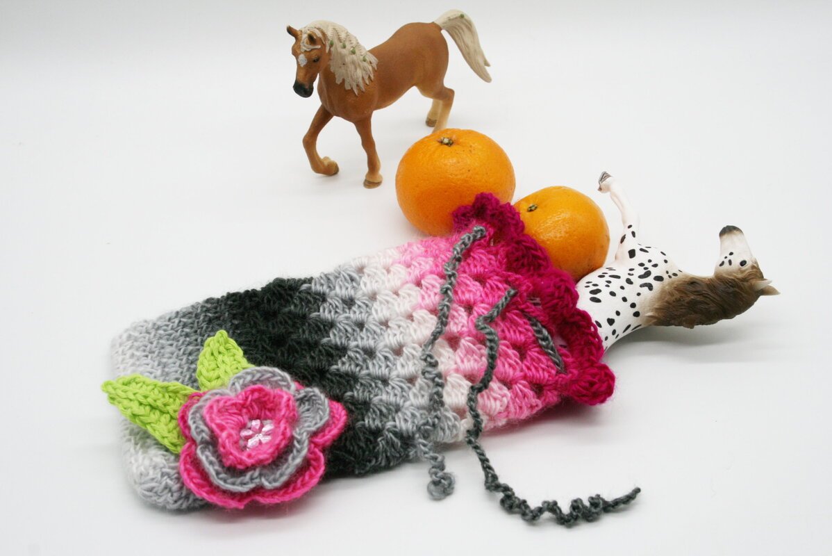 Gehäkelter Beutel in Grau‑ und Pinktönen mit mehrlagiger Blüte und Blättern, mit Zugband, liegend neben Orangen und Spielzeugpferden auf weißem Hintergrund.