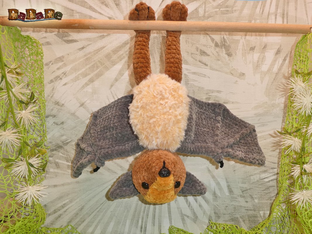 Gehäkelte Fledermaus in Braun, Beige und Grau hängt kopfüber an einer Holzstange vor hellem Blattmuster, seitlich grüne gehäkelte Vorhänge.