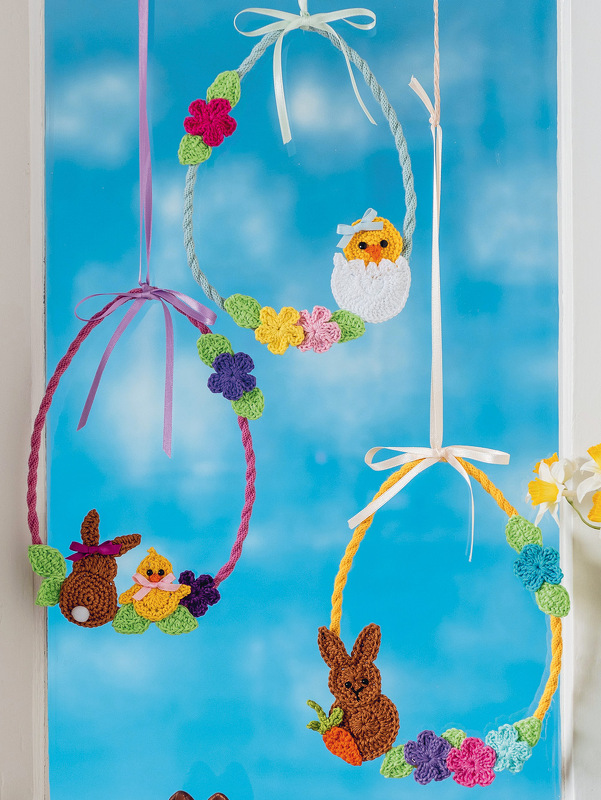 Drei handgehäkelte Osterkränze in verschiedenen Farben, geschmückt mit Hasen, Küken und Blumen, hängend vor einem Fenster.