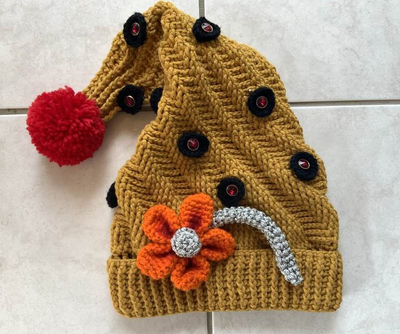 Gehäkelte senfgelbe Zipfelmütze mit rotem Pompon, schwarzen Knöpfen und oranger Blume