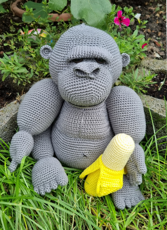 Gehäkelter grauer Amigurumi-Affe sitzt im Gras und hält eine gelbe gehäkelte Banane