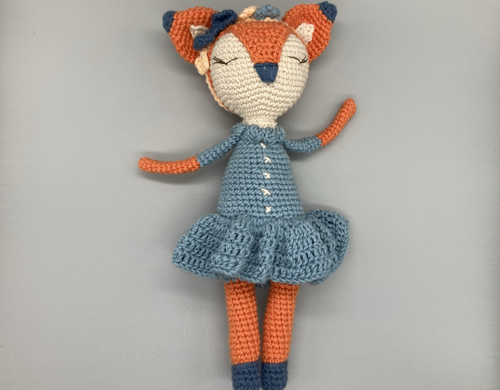 Gehäkelte Amigurumi-Fuchs-Puppe in hellblauem Kleid auf neutralem Hintergrund