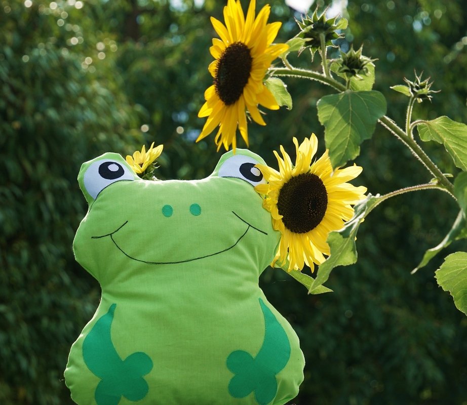 Grünes Frosch-Kuscheltier mit runden Augen und Lächeln vor Sonnenblumen