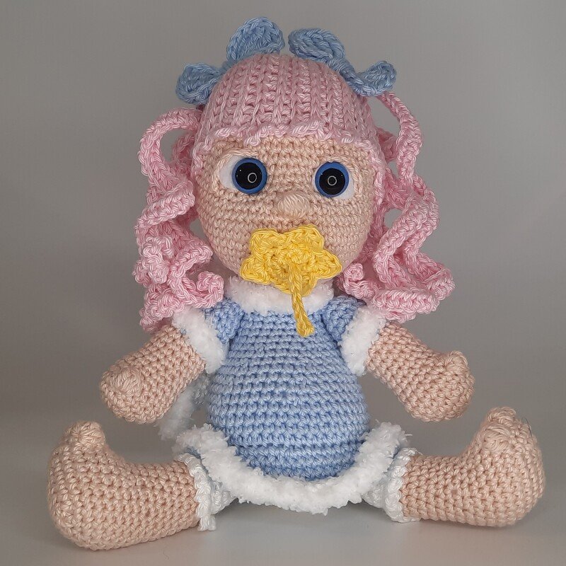 Sitzendes Amigurumi-Engelchen mit rosa Locken, hellblauem Kleid mit weißem Saum und gelbem Häkelstern vor dem Mund.