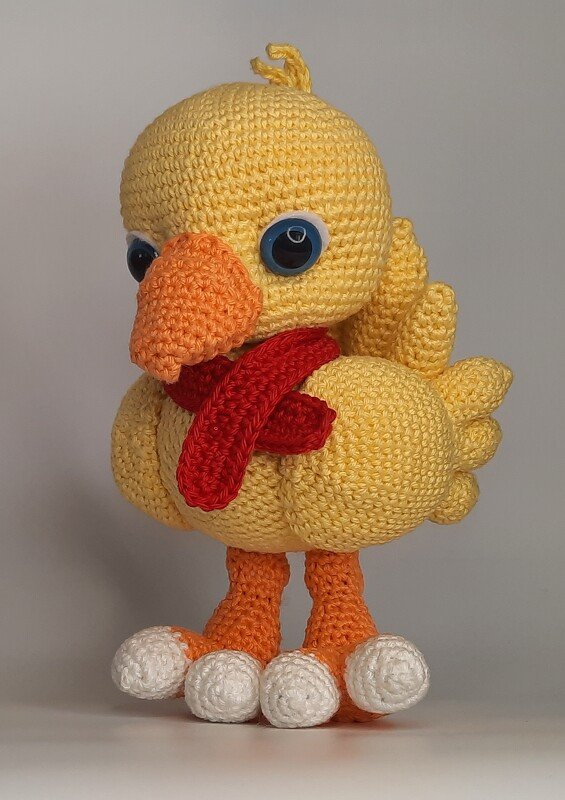 Gelbes gehäkeltes Amigurumi-Küken mit rotem Schal, orangem Schnabel und blauen Augen