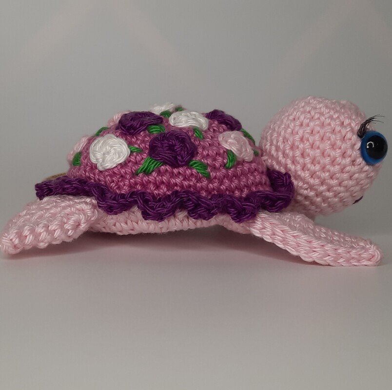 Seitliche Ansicht einer rosa gehäkelten Amigurumi-Schildkröte mit lila-blütenverziertem Panzer auf weißem Hintergrund