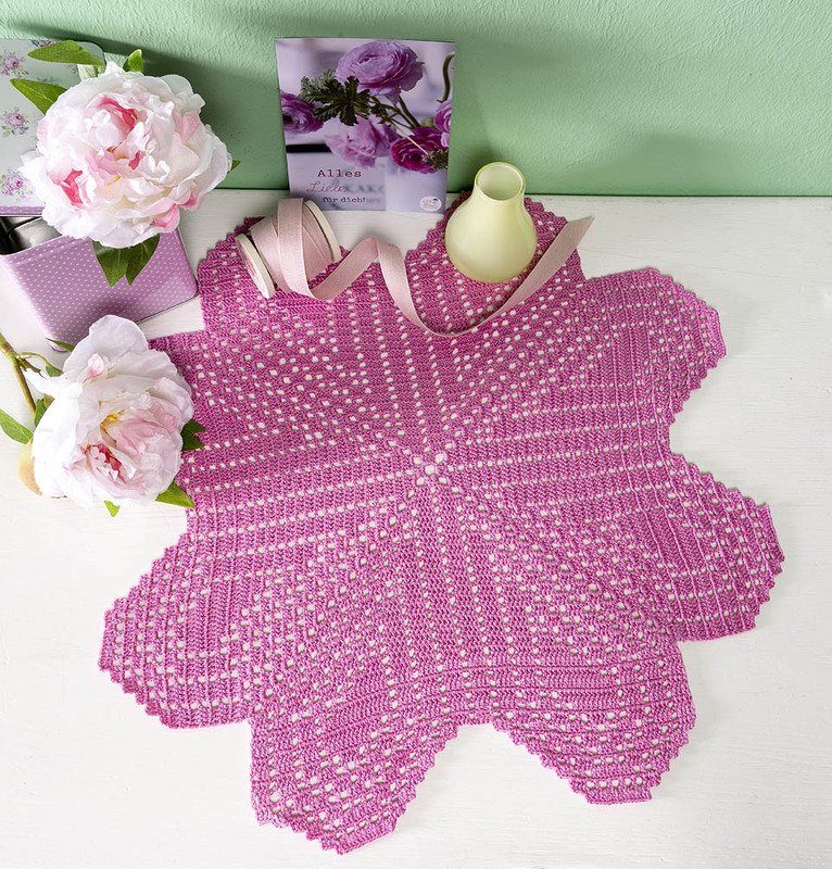 Magenta gehäkeltes Stern-Deckchen mit durchbrochenem Muster, neben rosa Pfingstrosen und einem Band auf hellem Untergrund.