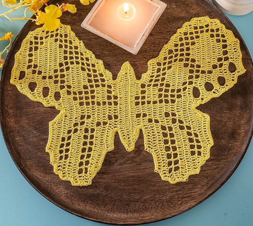 Gelbes gehäkeltes Schmetterlingsdeckchen auf dunklem Holztablett mit Kerze und gelben Blüten.