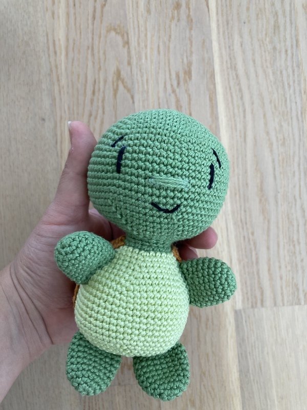 Handgefertigte grüne Amigurumi-Schildkröte mit hellem Bauch, von einer Hand gehalten vor hellem Holzboden.