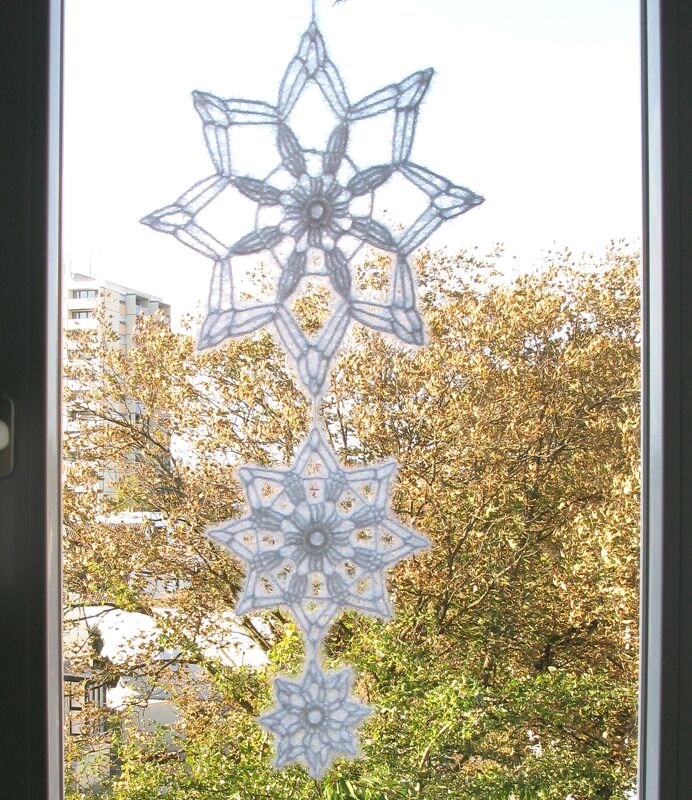 Drei weiße gehäkelte Schneeflocken als Kette hängend vor einem Fenster mit Bäumen im Hintergrund