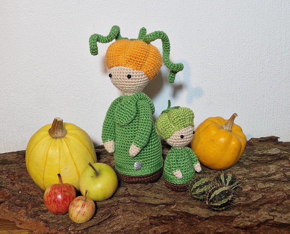 Zwei gehäkelte Amigurumi-Puppen im Kürbis-Design mit grünen Kleidern, arrangiert auf Baumrinde mit Äpfeln und Kürbissen.
