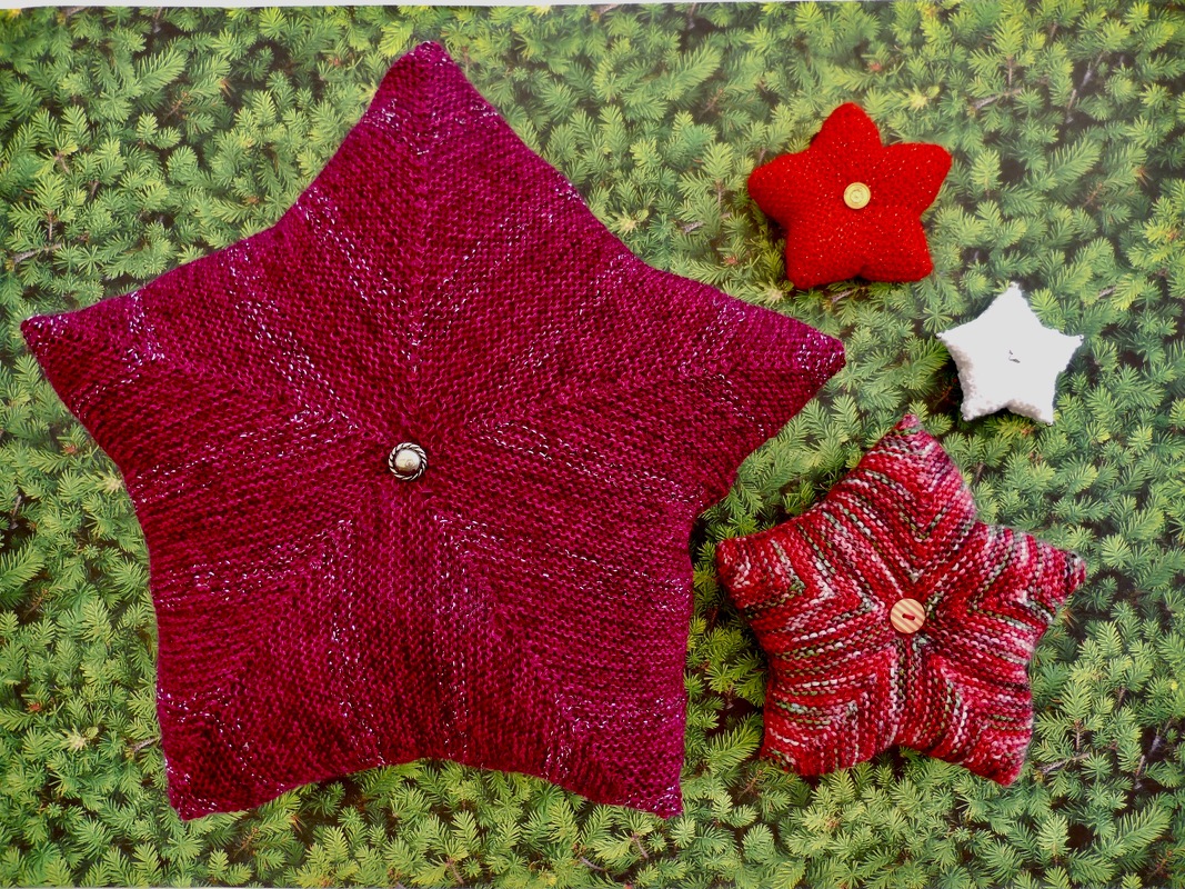 Vier gestrickte Sternkissen in Magenta, Rot, Rot-Weiß und Weiß auf grünem Nadelbaum-Hintergrund.