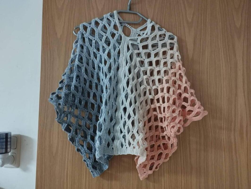 Gehäkelter Poncho mit Lochmuster in Blau–Grau–Rosa an einem Kleiderbügel vor einer Holztür