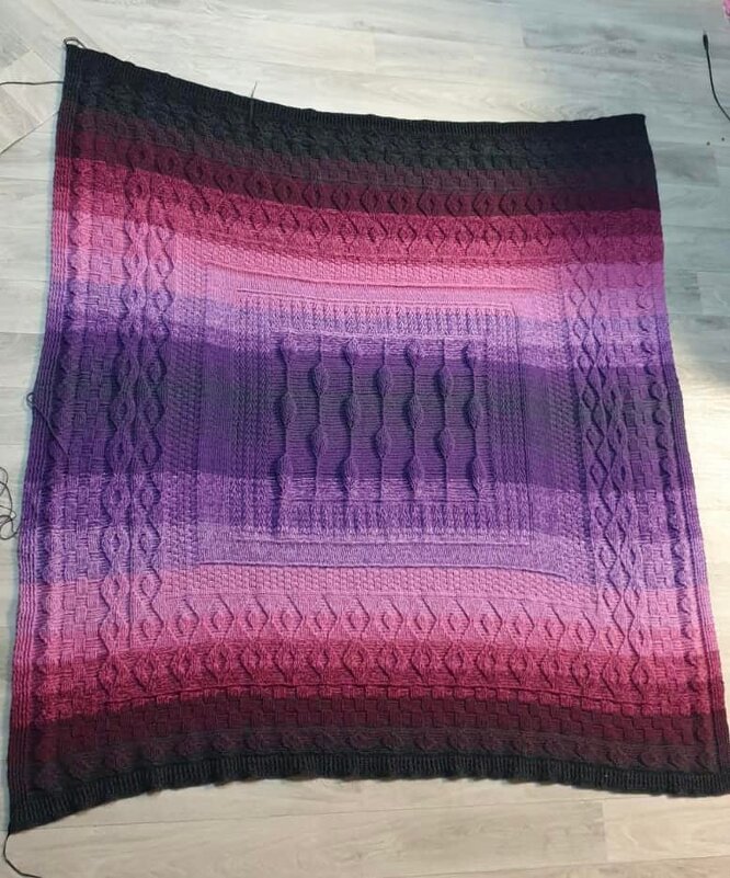 Rectangular crocheted blanket with an ombre gradient from dark charcoal to pink and purple, showing a central cable panel and textured stitch bands on a light wooden floor.