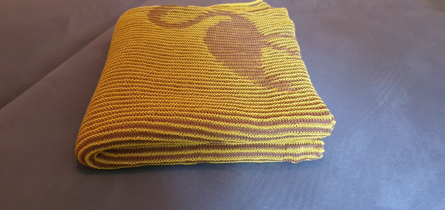 Folded yellow and brown knitted blanket with a large dark brown heart motif on a dark surface.