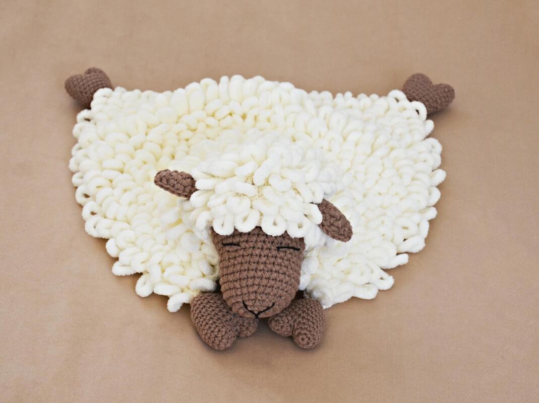 White looped yarn baby blanket shaped like a sleeping sheep with a brown crocheted head and feet on a neutral brown surface.
