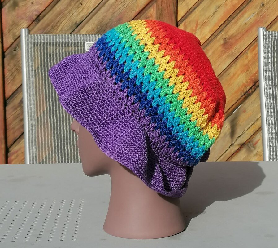 Seitliche Ansicht einer gehäkelten Damenmütze mit lila Schirm und regenbogenfarbenem Streifenmuster auf einem Schaufensterkopf
