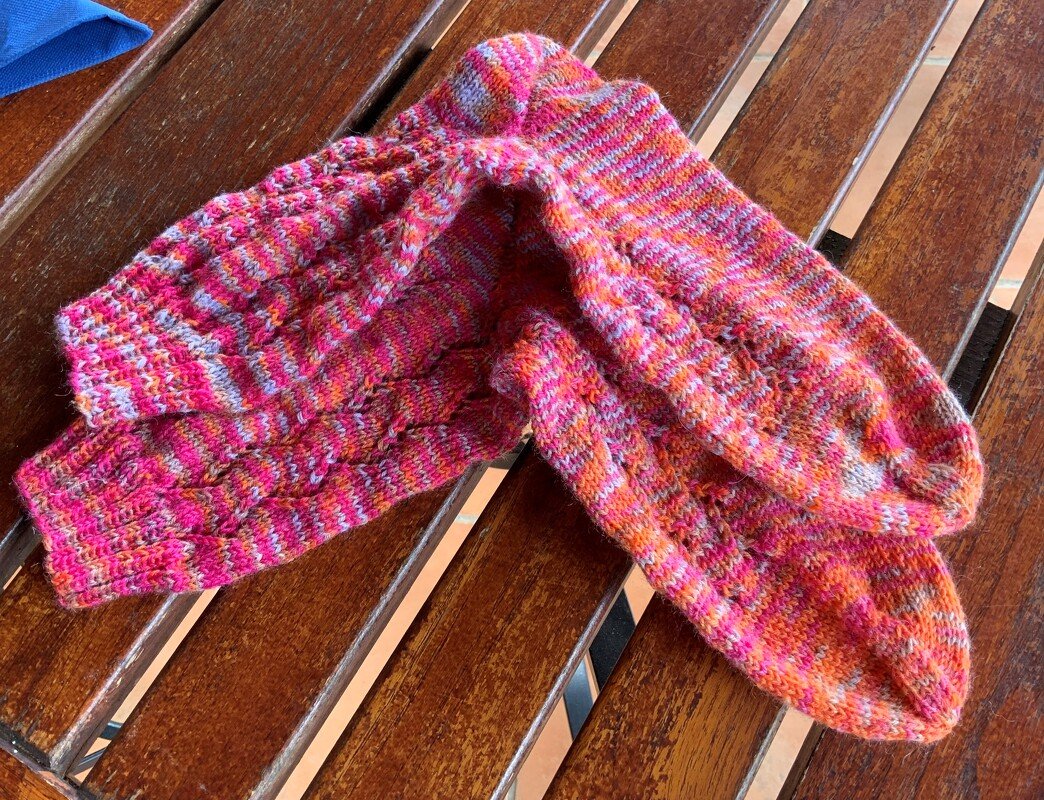 Handgestricktes Paar Socken in Pink, Orange und Grau mit feinem Lochmuster auf Holzoberfläche.