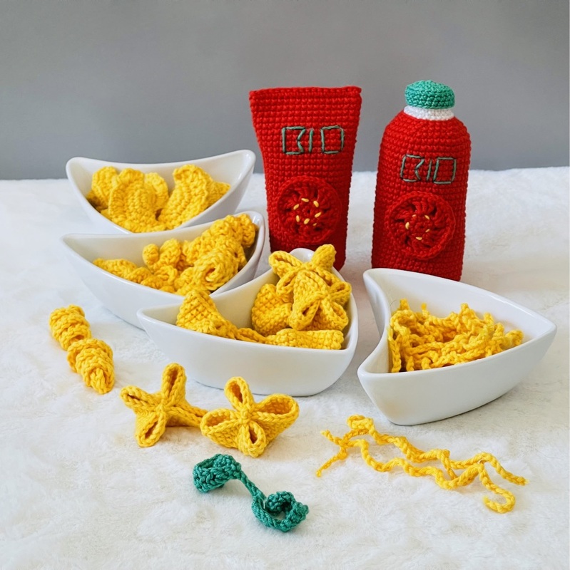 Gehäkeltes Spielzeugessen mit gelben Pommes- oder Nudelformen in weißen Schalen und zwei roten Flaschen