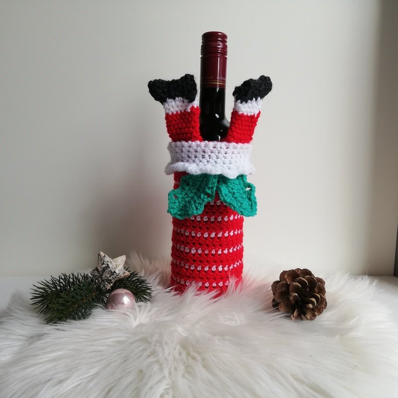 Flasche mit gehäkelter Weihnachts-Hülle und Santa-Beinen, daneben Tannenzweig und Zapfen