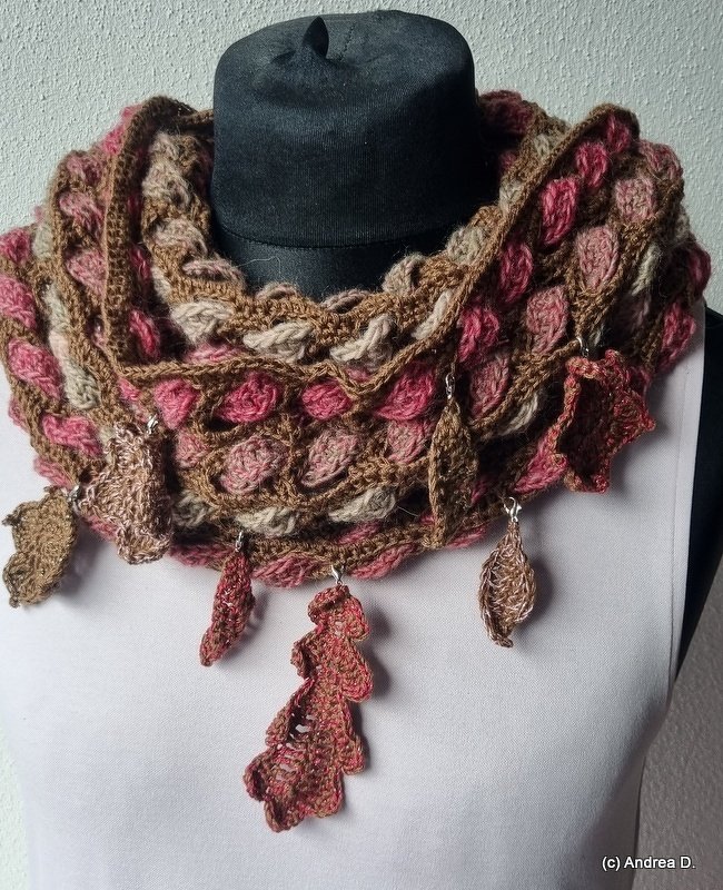 Mannequin wearing a brown, pink and beige crocheted loop cowl with textured loop stitch and small crocheted leaf dangles along the lower edge.