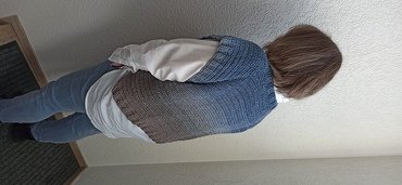 Rückenansicht einer Person in gehäkelter Weste mit blau-braunem Farbverlauf über weißem Oberteil und Jeans.