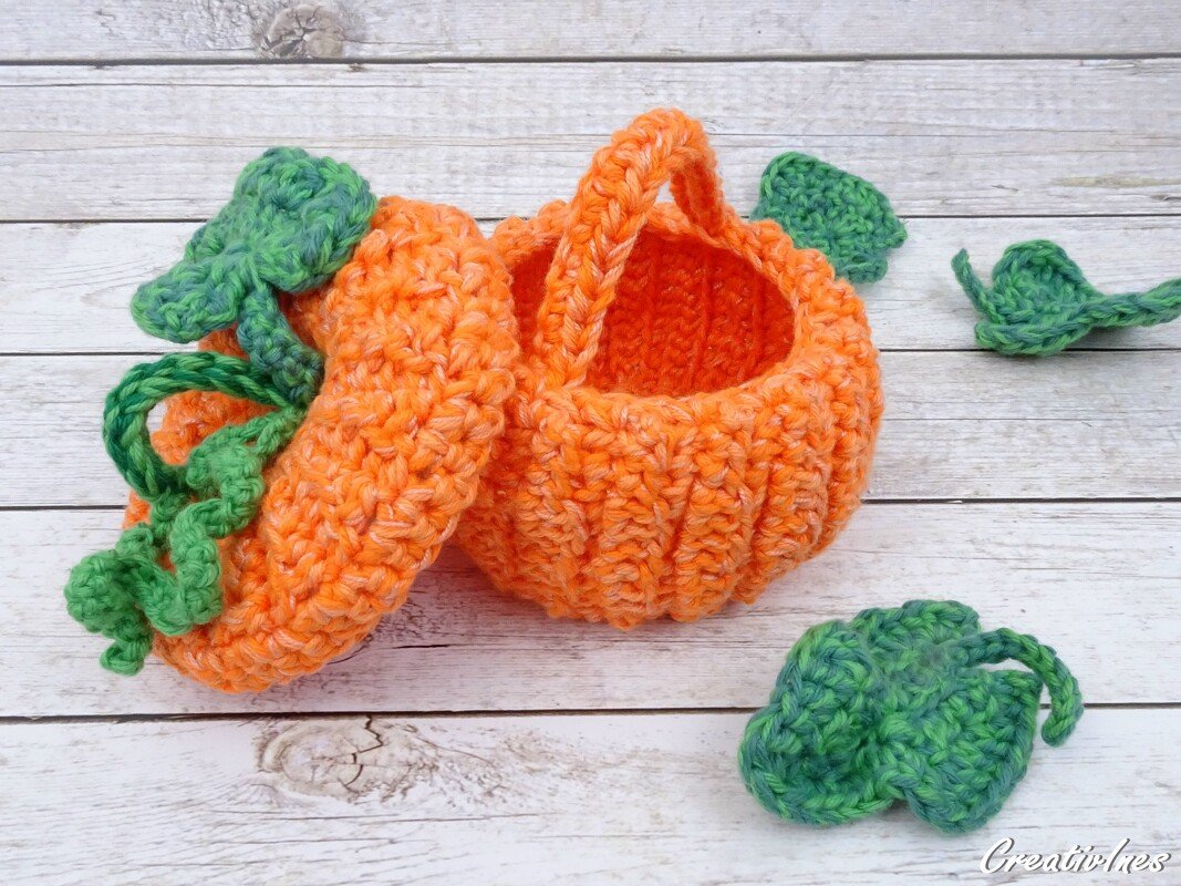 Gehäkelter orangefarbener Kürbiskorb mit Deckel, Henkel und grünen Blättern auf heller Holzfläche