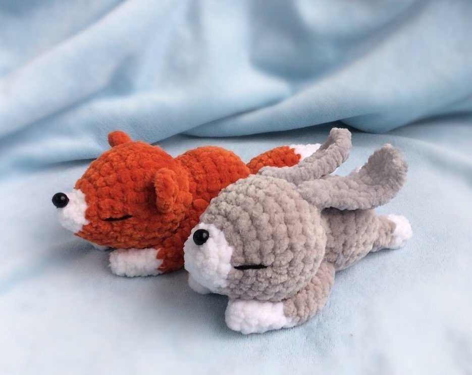 Oranger Amigurumi-Fuchs und grauer Amigurumi-Hase liegen nebeneinander auf hellblauem Stoff.