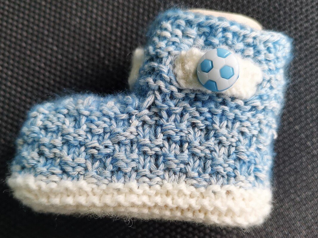 Gestrickter blau-weiß melierter Babyschuh mit weißer Kante und Fußballknopf auf dunklem Hintergrund
