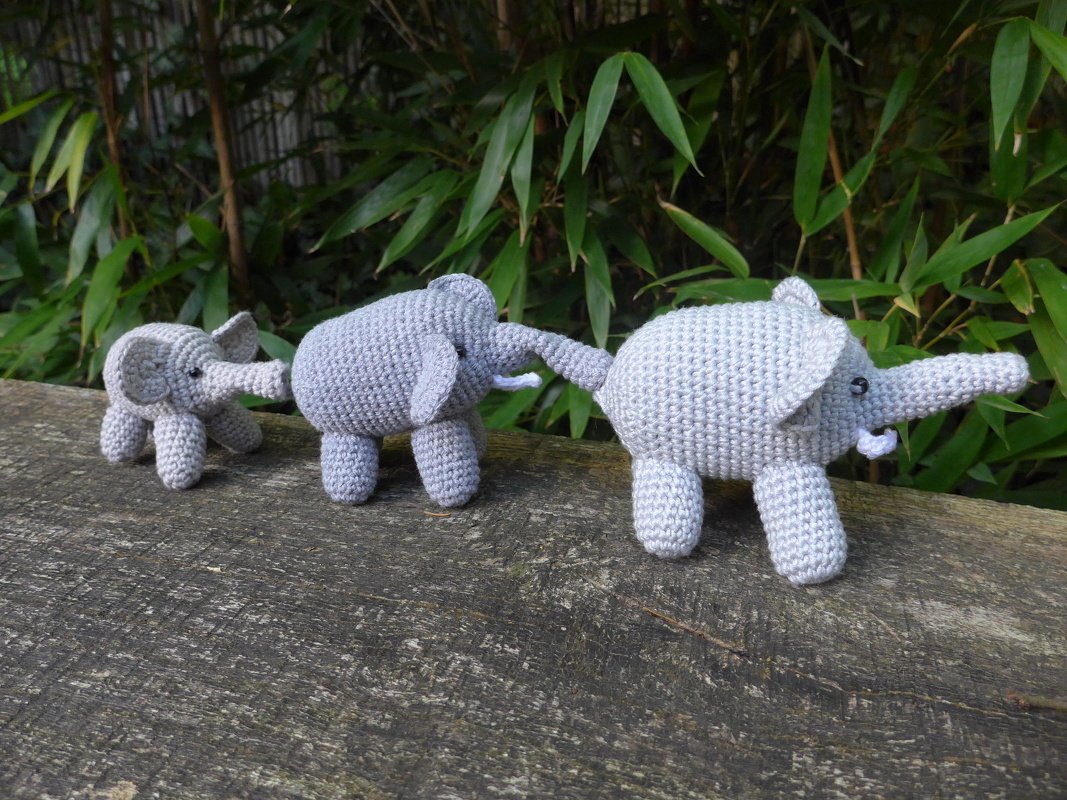 Drei gehäkelte Elefanten-Amigurumi in Grautönen auf einer Holzoberfläche, mit grünen Pflanzen im Hintergrund.