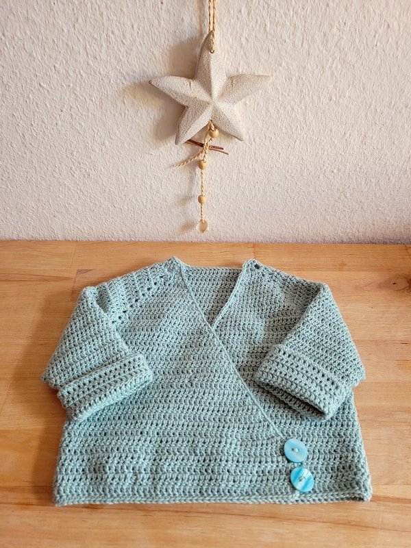 Gehäkelte mintgrüne Baby-Wickeljacke mit zwei türkisfarbenen Knöpfen auf Holz, Stern-Deko an weißer Wand