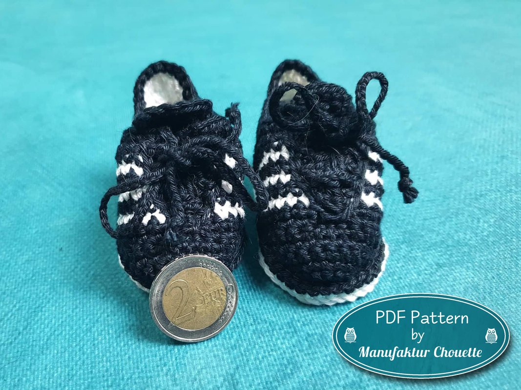 Vorderansicht zweier handgehäkelter Mini‑Sneaker in Schwarz‑Weiß neben einer 2‑Euro‑Münze zum Größenvergleich