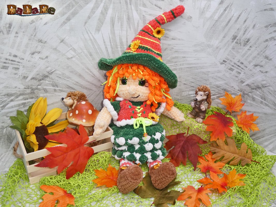 Gehäkelte Amigurumi-Hexe mit grün-rotem Spitzhut, orangefarbenen Haaren, Sonnenblume und Herbstblättern, umgeben von Igel- und Pilzfiguren.