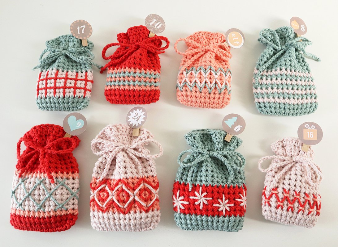 Acht gehäkelte Adventsbeutel in Pastellgrün, Rosa und Rot mit Zugschnüren und nummerierten Holzklammern auf hellem Hintergrund