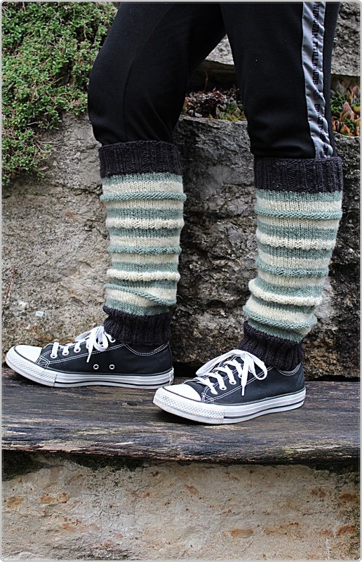 Gestrickte, gestreifte Beinstulpen in Dunkelgrau, Cremeweiß und Grünblau, getragen über dunklen Hosen mit schwarzen Turnschuhen.
