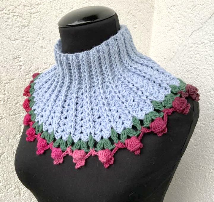 Romantischer hellblauer Häkelkragen mit pink-roter Blütenborte und grünen Blättern auf dunklem Mannequin