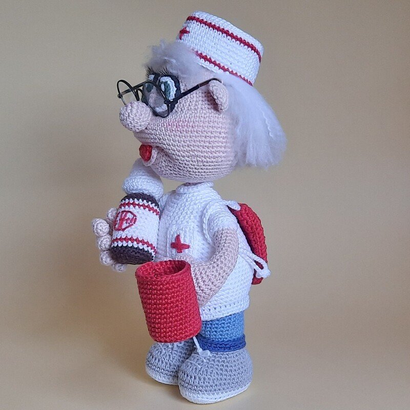 Gehäkelter Krankenpfleger als Amigurumi im Profil mit rotem Rucksack, Flasche und rotem Becher vor neutralem Hintergrund.