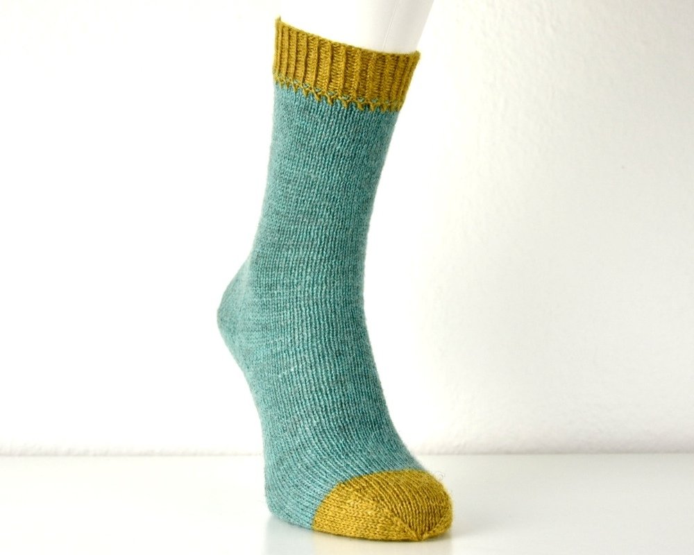 Gestrickte Socke in Türkis mit senfgelbem Bündchen und Spitze auf weißem Hintergrund.