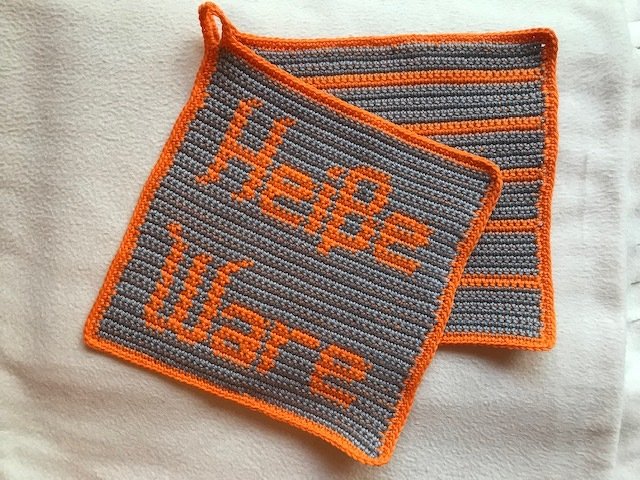 Zwei gehäkelte Topflappen in Grau und Orange; einer mit Schrift 'Heiße Ware', der andere gestreift