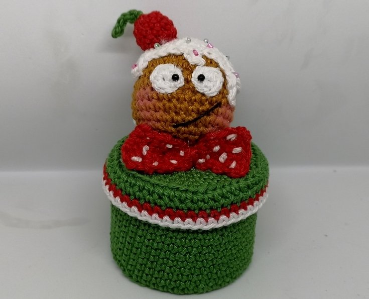 Amigurumi-Figur mit roter Fliege, weißem Guss und roter Kirsche auf dem Kopf, die auf einer grünen Häkelbox sitzt