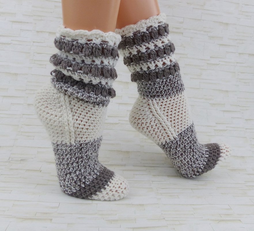 Nahaufnahme zweier gehäkelter Socken in Cremeweiß und Graubraun mit Blockstreifen und gerafftem Schaft an Modellbeinen vor hellem Hintergrund.