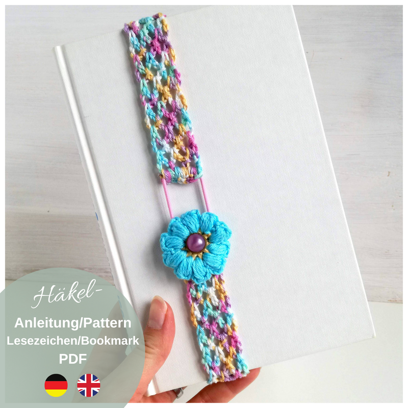 Hand holding white hardcover book with a multicolored crocheted strap bookmark and a turquoise flower attached by an elastic loop