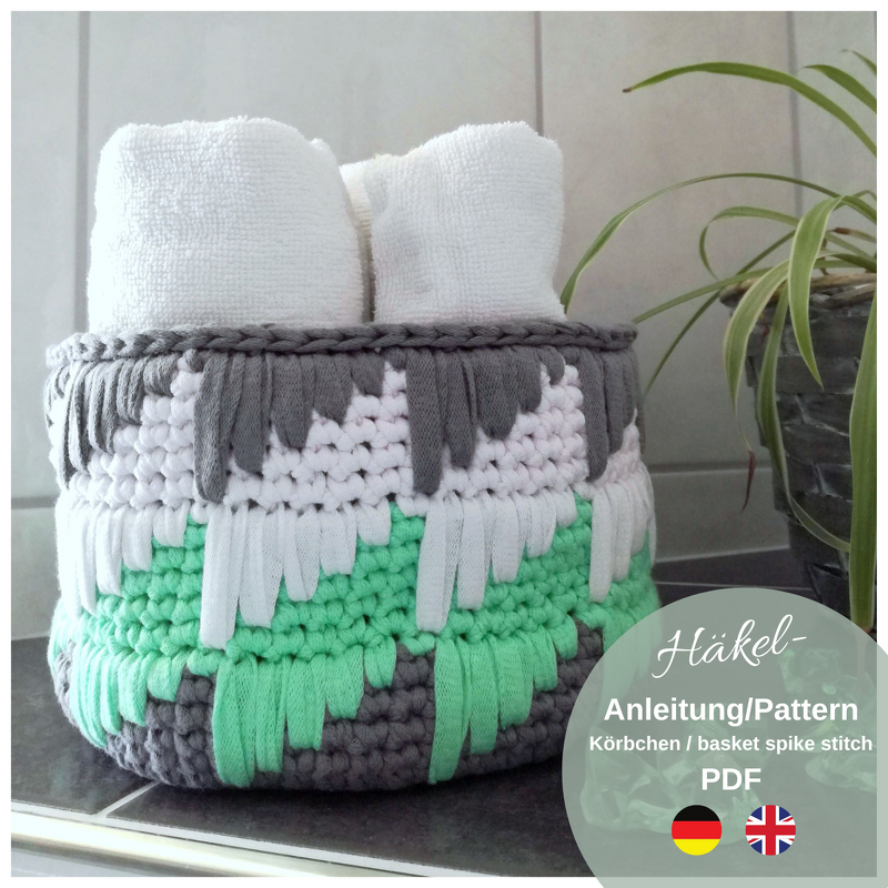 Round crochet basket in gray, white and mint with spike-stitch fringe, holding two rolled white towels on a tiled bathroom surface beside a potted plant