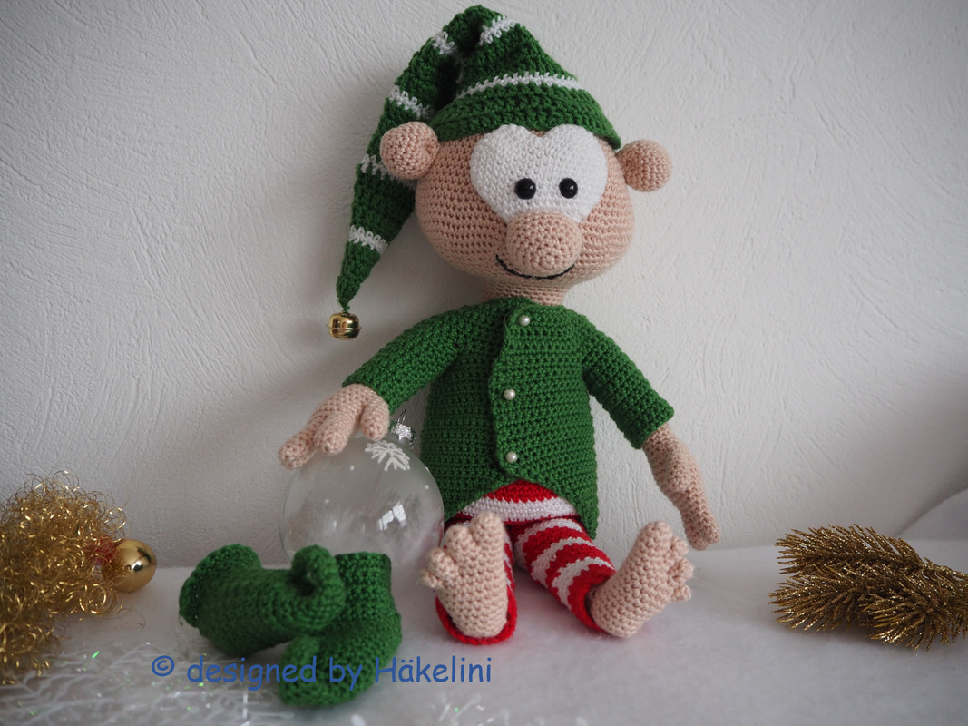 Gehäkelter Weihnachtself in Grün, Rot und Weiß mit Zipfelmütze, Jacke und gestreiften Hosen, umgeben von Weihnachtsdekoration.