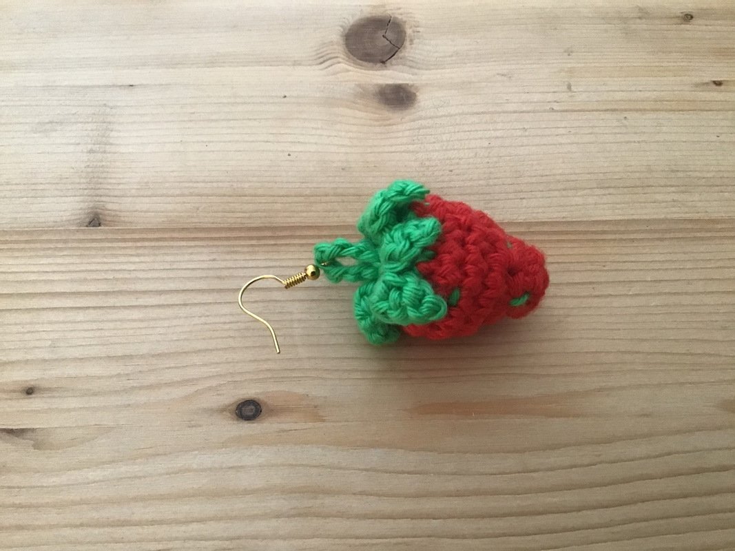 Roter gehäkelter Erdbeer-Ohrring mit grünen Blättern und goldfarbenem Haken auf Holzuntergrund.