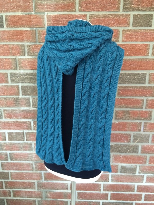 Petrolfarbener gestrickter Kapuzenschal mit Zopfmuster auf einer Schneiderpuppe vor Backsteinwand.